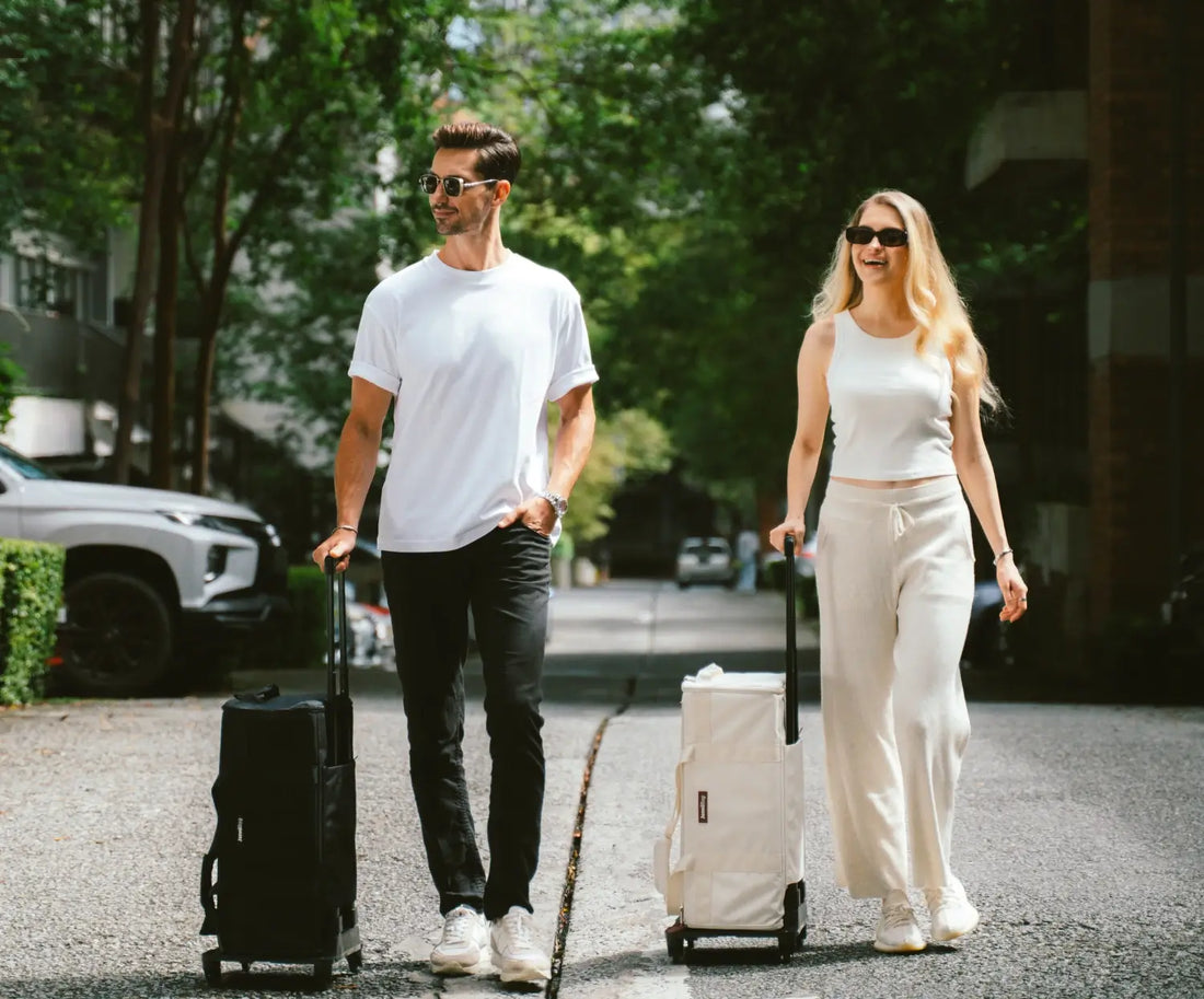 Man and woman walking with suitcases on a path surrounded by trees.